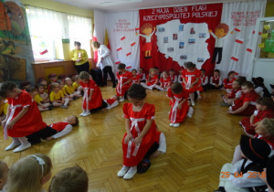 Na tle dekoracji z mapą Polski i dziećmi z chorągiewkami przedszkolaki wykonują układ taneczny do utworu "Prząśniczki".